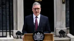 ダウニング街の首相官邸の前で、マイクの載った演台に向かい話すダークスーツ姿のスターマー氏