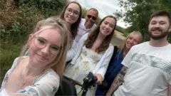 Foto de Megan com a família. Ela está na frente, sentada em uma cadeira de rodas. Há outras cinco pessoas na foto, sendo três mulheres e dois jovens.