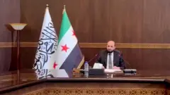 Костюм-шим кийган эркак орқасида катта байроқлар билан стол бошида ўтирибди