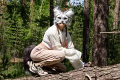 A teenage girl wearing an animal face mask and bushy fake tail crouches on a fallen tree looking into the camera.