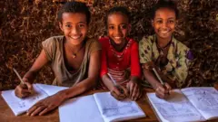 ክልተ ደቀንስትዮን ሓደ ወዲ ተባዕታይን ተምሃሮ ብፍሽኽታ ናብ ካሜራ እናጠመቱ፡ ኣብ ልዕሊ ዝተገንጸለ ጥራውዝቶም ቢሮኦም ሒዞም ይርኣዩ።