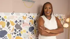 Deidre Mathis sorrindo com um vestido branco, em frente a uma parede coberta de papel estampado, com um tecido de fundo rosa e os dizeres 'Wanderstay'