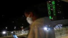 Foto à noite, mostra mulher parada em ponto de ônibus com máscara e, ao fundo, uma projeção de luz no prédio do Congresso dizendo: Luto 100 mil