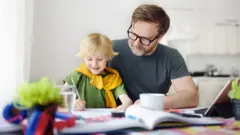 Ojciec pomaga synowi w pracy domowej. Obaj siedzą przy biurku w jasnym pokoju o białych ścianach. Na pierwszym planie leżące na biurku książki i pomoce do nauki. 