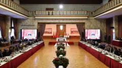 Kurtulmuş'un ana kürsüde oturduğu salonda sağda ve solda uzun kırmızı etek beyaz örtülü masalarda milletvekilleri oturuyor. Orada kırmızı çiçekler sıralanıyor. 