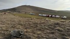 Uçağın enkazından bir parça bir tarlada görünüyor ve arka plandaki tepenin alt kısmında araçlar park etmiş halde duruyor