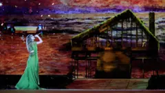Taylor Swift no palco, usando um vestido longo verde enquanto canta diante de um cenário que reproduz uma cabana de madeira coberta de vegetação. Ao fundo, um grande painel exibe uma paisagem em tons alaranjados e arroxeados.