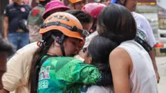 အစိမ်းရောင် ရှပ် ဝတ်ထားပြီး လိမ္မော်ရောင် ဦးထုပ်ဆောင်းထားတဲ့အမျိုးသမီးက သူ့လက်ထဲမှာ ပွေ့ထားတဲ့အမျိုးသမီးကို နှစ်သိမ့်ပေးနေတာဖြစ်ပါတယ်။ သူ့နောက်မှာ ရှာဖွေ ကယ်ဆယ်ရေးသမားတွေ ရှိနေ 