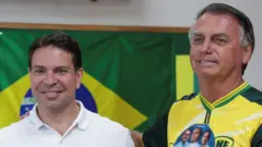 Alexandre Ramagem aparece à esquerda, usando uma camisa polo branca e sorrindo, enquanto Jair Bolsonaro está à direita, vestindo uma camiseta amarela com detalhes em verde e um adesivo com rostos, também sorrindo. Bolsonaro apoia o braço no ombro de Ramagem, e ao fundo há uma bandeira do Brasil.