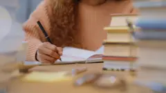 Jovem estudando, escrevendo em um caderno, rodeada por vários livros