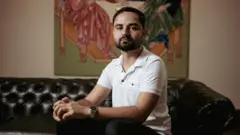 Kaik Ribas sentado num sofá preto, com uma pintura colorida ao fundo