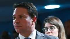 Alexandre Ramagem aparece em primeiro plano, usando terno escuro, camisa branca e gravata, com expressão séria e olhando para o lado. Ao fundo, ligeiramente desfocada, há uma mulher de óculos e cabelos longos, também com expressão neutra.