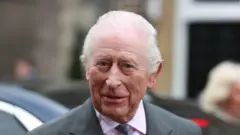 King Charles III smiles at the crowd during a visit to Banbridge Old Town Hall on day three of the royal visit to Northern Ireland - he has a slight smile, and is wearing a gray suit, with a pinkish shirt and tie 