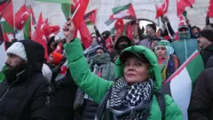 Yeşil montlu bir kadın elinde bayrakla duruyor. Etrafında soğuktan korunmak için sıkı giyinmiş çok sayıda kişi var.