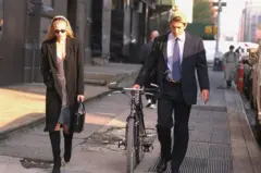 Carolyn Bessette y John F. Kennedy caminan por una calle. Ella lleva unos lentes de sol, viste unas botas largas negras, un vestido corto y un sobretodo negro. El tiene una gorra de beisbol volteada hacia atrás, viste un traje negro, con camisa azul y corbata y arrastra con su mano derecha una bicicleta.