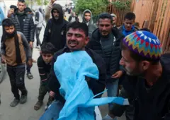 الأب بشعره الأسود وبشرته السمراء، يحمل جثمان ابنه الملفوف بقماش أرزق، وحوله عدد من الرجال والأطفال الفلسطينيين. الصورة التقطت نهاراً.