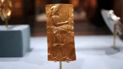 Placa dourada mostrando um homem de perfil com um objeto longo na mão. Ele veste um gorro, uma túnica curta e calças, com uma espécie de espada na cintura.