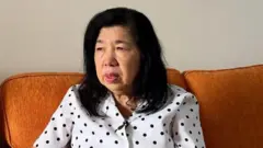 Susanna Liew, vestida con un top blanco con lunares negros, está sentada en un sofá marrón.