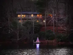 Una casa de dos plantas frente al lago, rodeada de altos árboles, con un árbol de Navidad iluminado. La luz cálida sale a través de las ventanas y las luces del árbol se reflejan en la superficie del lago.