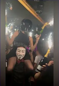 Una persona con un casco de moto está sentada sobre los hombros de otra persona. Ambas sostienen botellas de champán con bengalas encendidas, cerca del techo, mientras caminan por un bar abarrotado de gente.