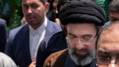 Mojtaba Khamenei, caminhando ao lado de outros homens, vestido com roupas tradicionais com barba grisalha, óculos e olhando para baixo
