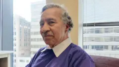 Justino Gómez, un hombre mayor que viste una camisa blanca y un suéter morado, sentado con edificios de gran altura detrás de él.