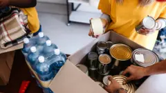 Pessoas com camisetas amarelas colocam alimentos enlatados dentro de uma caixa de papelão para doação