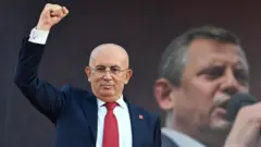 Ümit Erkol sağ yumruğun havaya kaldırmış halde görülüyor. Üzerinde koyu lacivert bir takım elbise ve kırmızı kravat var. 