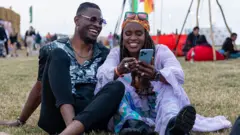 Două persoane stau pe iarbă la un festival, amândouă zâmbesc, una ține un telefon și se uită împreună la ecran, cu alte persoane în jurul lor.