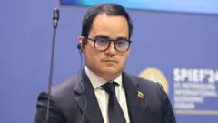 Calixto Ortega Sánchez, vestido de terno e gravata, com um pin com a bandeira da Venezuela e fone de ouvido