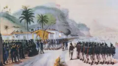 A imagem mostra uma cerimônia militar à beira-mar, com uma tropa de soldados em formação diante de autoridades e civis reunidos em frente a casas coloniais. Ao fundo, destacam-se morros cobertos por vegetação, palmeiras e embarcações ancoradas na baía, compondo uma cena de caráter histórico.