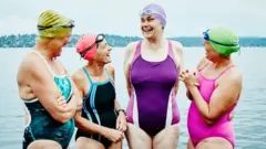 Cuatro alegres mujeres de mediana edad en vestidos de baño frente a un lago