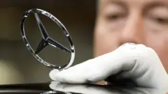 An employee wearing white gloves attaches the Mercedes-Benz star ornament to the front of an S-Class car