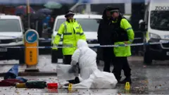 Dois policiais com coletes de alta visibilidade e outra pessoa com casaco preto com capuz estão na rua. Uma pessoa com macacão branco forense está ajoelhada à frente deles. Ao fundo, há viaturas da polícia e equipamentos forenses espalhados no chão.
