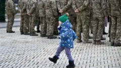 Дитина у синій куртці і зеленій шапці йде вулицею. Поруч стоять військові. 