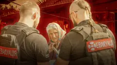 Двоє чоловіків у бронежилетах із написом «ЕВАКУАЦІЯ» спілкуються з літньою жінкою, яка тримає документи та стривожено дивиться повз камеру. Сцена виглядає як допомога цивільним у зоні небезпеки.
