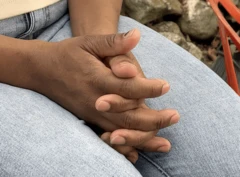 Mãos de Paula sobre as pernas usando calça jeans e uma camisa creme
