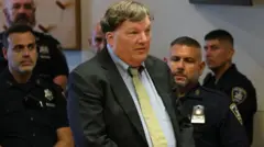Rex Heuermann, con traje oscuro y camisa y corbata claras en el tribunal, con las manos entrelazadas y gesto serio, rodeado de agentes de policía y una mujer sentada a su derecha