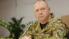 Військовослужбовець у пікселі ЗСУ сидить за столом у приміщенні. На його формі видно шеврон із українським прапором та інші службові нашивки.
