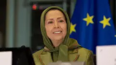 Maryam Rajavi com um lenço verde cobrindo a cabeça e uma jaqueta da mesma cor, em frente à bandeira da União Europeia