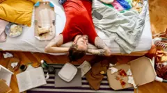 লাল টিশার্ট ও নীল-সাদা চেক ট্রাউজার পরা একজন ব্যক্তি বিছানার ওপর মাথার পেছনে দুই হাতের তালু রেখে শুয়ে আছেন