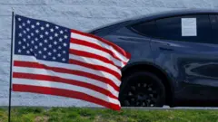 加州爾灣某二手車商賣場外一輛特斯拉 Model Y 停放在一面美國國旗旁（27/3/2025）