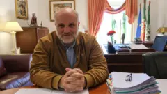 Paulo Fernandes, um homem calvo de meia idade, sentado em sua mesa de trabalho coberta de papéis, com as mãos cruzadas e vestindo um casaco bege, sobre camisa azul clara