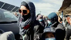 A woman in a headscarf and dark glasses with other women in headscarves and faces masks behind her.