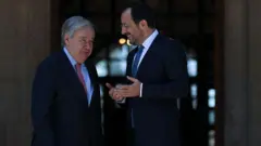 Beyaz saçlı gülen bir takım elbiseli erkek ile çok daha genç takım elbiseli bir diğer siyasetçi ellerini açmış şekilde ona yine bir şeyler anlatıyor. ikisi de bir kapı girişinde ayaktalar.