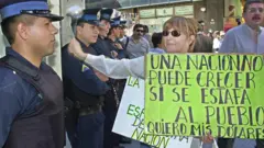 Uma mulher segurando cartaz com os dizeres: 'Uma nação não pode crescer enquanto as pessoas são enganadas. Quero meus dólares', enquanto bate na porta de um banco vigiado pela polícia em Buenos Aires, em abril de 2002