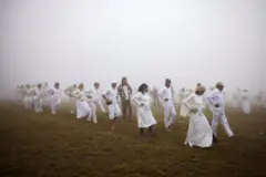 desetine ljudi obuceni u belu odecu izvode plesni ritual na planini okruzeni maglom
