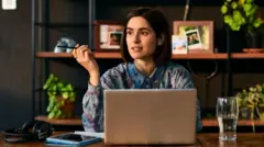 Bir kadın düşünceli bir şekilde laptop başında oturuyor, ofise benzer bir alanda masasının üzerinde bardakta su, kulaklıklar ve tableti var. Arka planda fotoğraf ve çiçeklerin olduğu bir raf. 