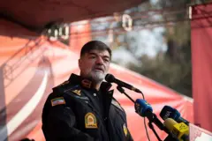 Em frente a um fundo vermelho, em um palco em ambiente externo, o brigadeiro-general Ahmad-Reza Radan fala para os microfones no dia 24 de novembro de 2025