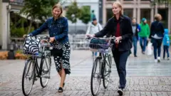 Duas mulheres caminham sorridentes, empurrando bicicletas em um calçadão de Oslo, na Noruega